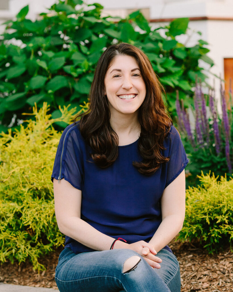 Woman with brown hair wearing a blue shirt with greenery in the background
