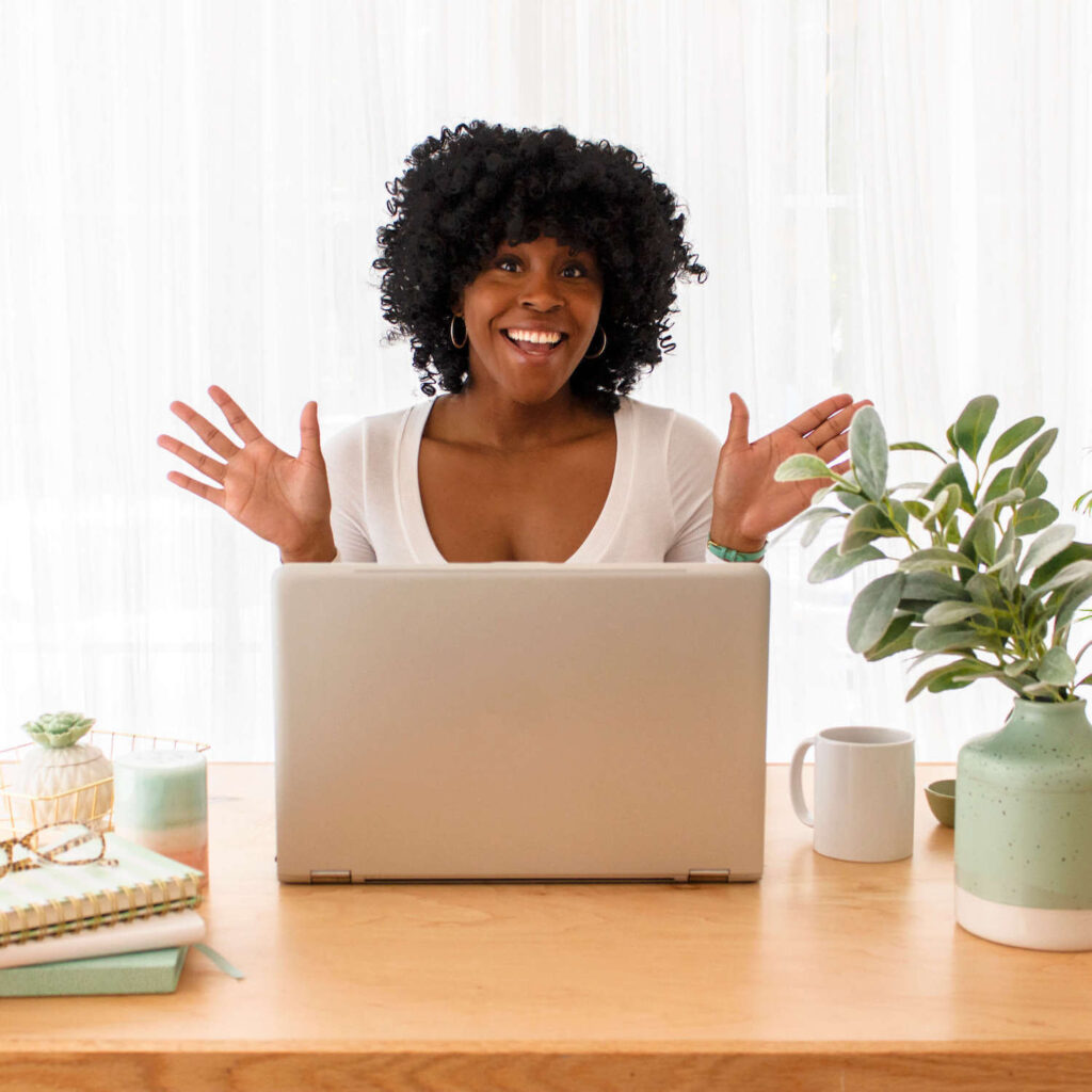 Woman sitting behind laptop looking at the camera smiling with hands in the air