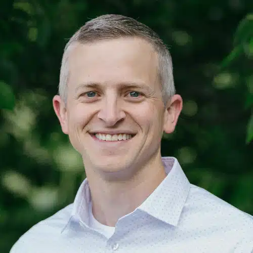 Image of Garret, a white man wearing a crisp white collard shirt, looking and smiling at the camera, dappled leaves in the background.
