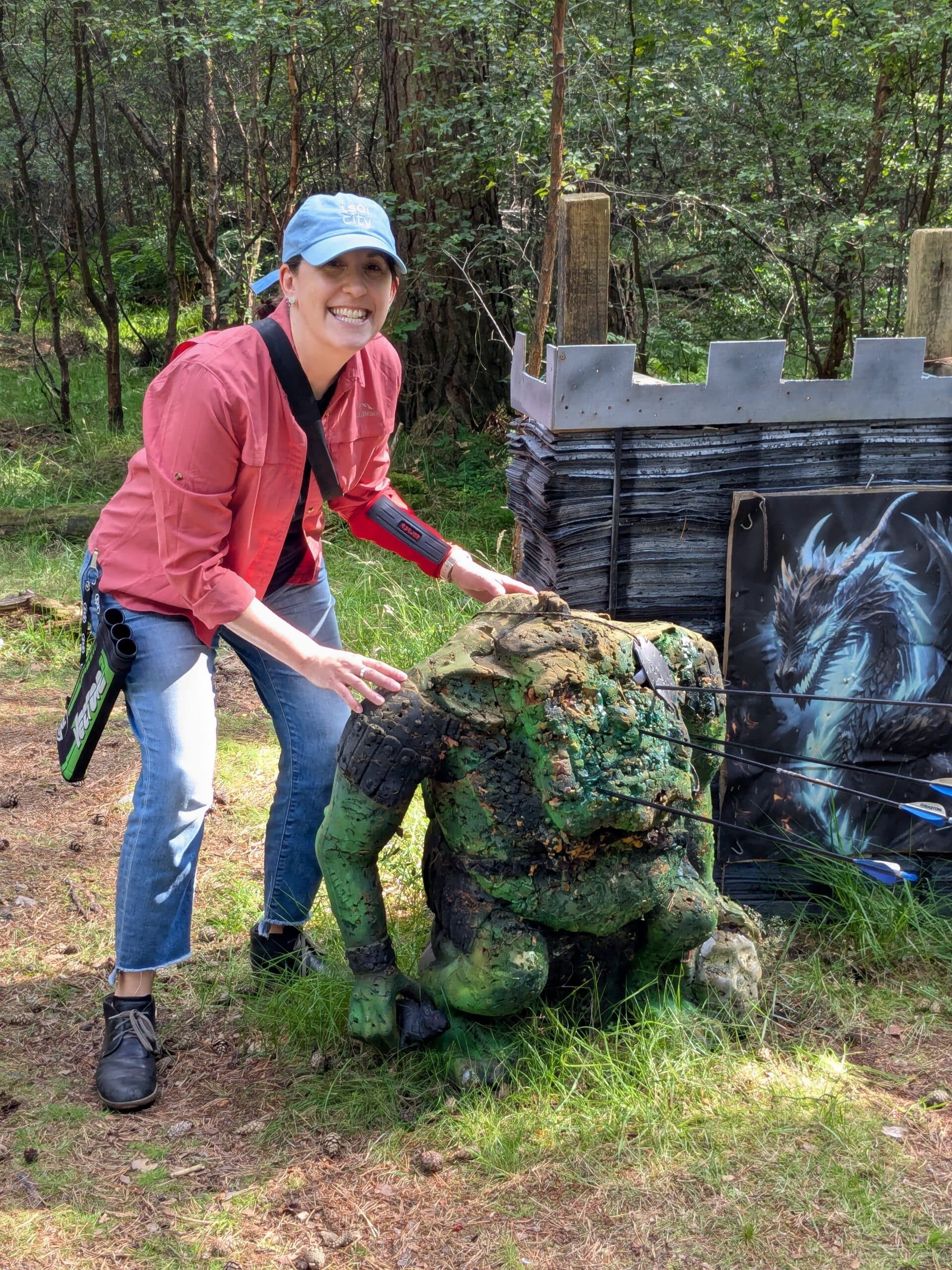 Bev Feldman in a forest archery course proudly standing next to a pretend ogre that she shot in the face with four arrows.